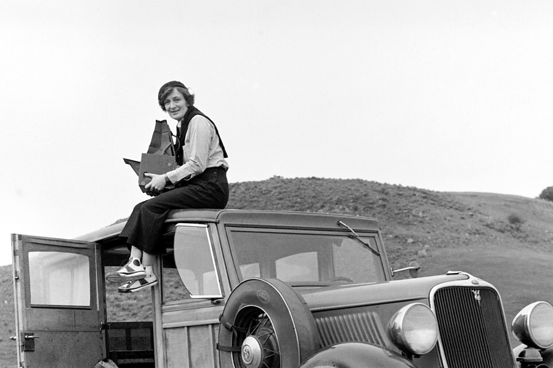 Photographer Dorothea Lange sitting on the roof of her 1933 Ford station wagon at the side of a California road in 1936, holding a large-format camera, the car door open beside her and a bare hillside behind.