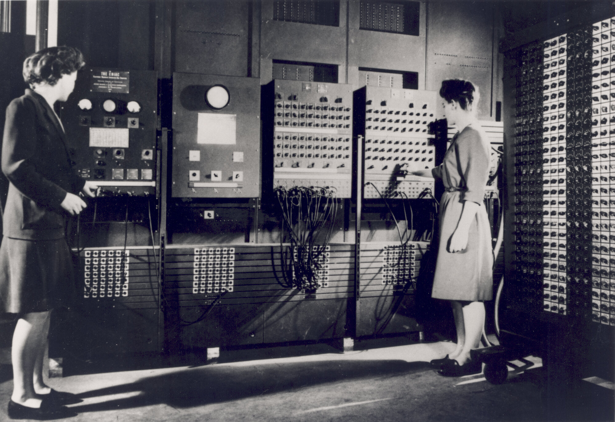 Two women operating the ENIAC computer around 1946: one stands at the left-hand control panel while the other adjusts a bank of switches and patch cables on the machine, in a room lined with equipment racks.