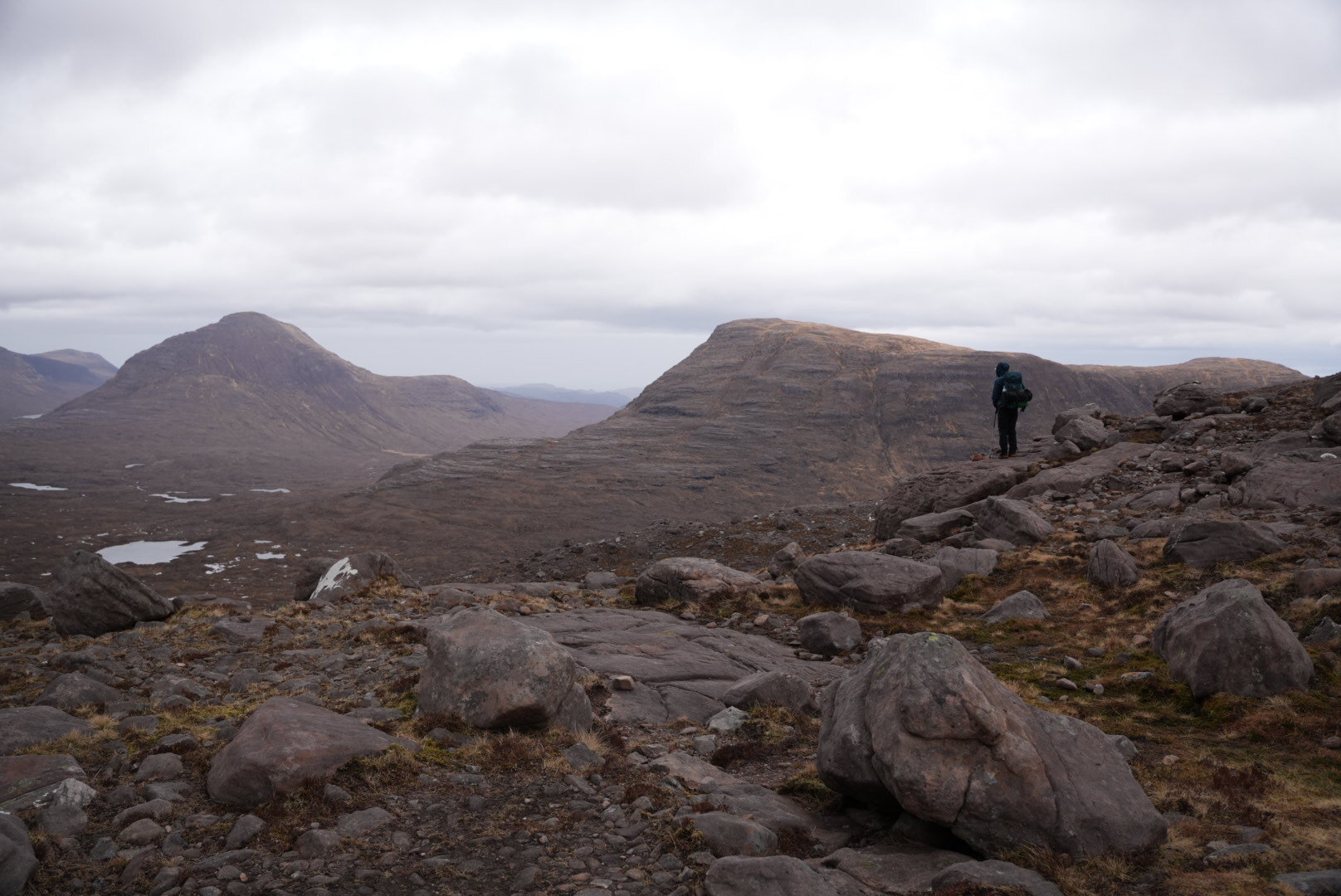 A lone hiker with a backpack stands on a boulder-strewn ridge, looking out over the brown hills and small lochans of the Scottish Highlands under a heavy grey sky.